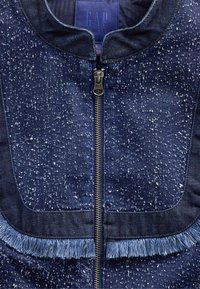 Jeans jakna s temno modrim pikčastim vzorcem, zadrgo za zapiranje in svetlo modrimi cofki ob robu in žepih. Vidna je blagovna znamka GAP.