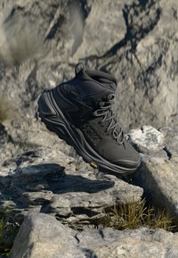 Svart vandringskänga med hög ankel, texturerat tyg, vadderad krage och Vibram-sula, som visas svävande över stenig terräng.