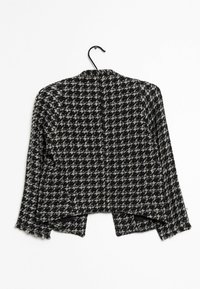 Czarna i biała kurtka w wzór houndstooth o skróconym kroju, postrzępionych krawędziach i otwartym przodzie, wisząca na czarnym haczyku.