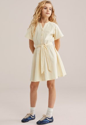 Mlada deklica z dolgimi kodrastimi lasmi, oblečena v bledo rumeno obleko z gumbi in pasom ter temno modre superge z belimi nogavicami.