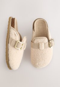 Beżowe, puszyste kapcie z teksturowanym wzorem i paskiem na klamrę. Miękki materiał i wkładka korkowa. Gumowa podeszwa zapewniająca przyczepność.