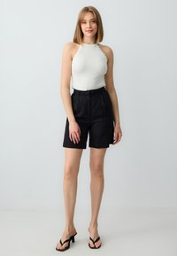 Shorts neri a vita alta con pieghe abbinati a un top a halter bianco aderente. Texture liscia e design minimalista. Sandali neri con tacco.