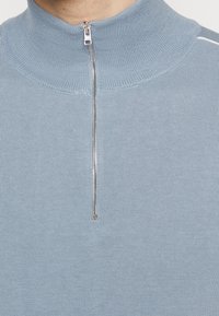 Ljusblå stickad tröja med hög krage och halvdragkedja framtill. Har ett strukturerat tyg utan mönster eller ytterligare detaljer.