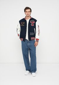 Chaqueta varsity en azul marino y blanca con texto bordado, acentos a rayas y logotipo; combinada con jeans azules con estampados y zapatillas blancas.