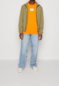 Hombre con chaqueta verde claro con cremallera, camiseta naranja brillante, pantalones de mezclilla de pierna ancha azul claro y zapatillas blancas contra un fondo plano.