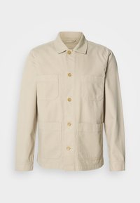 Beżowa bawełniana kurtka z zapięciem na guziki z przodu, dwoma kieszeniami na piersi i dwoma bocznymi kieszeniami. Posiada klasyczny kołnierz oraz długie rękawy.