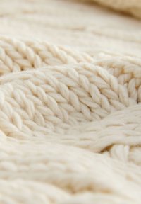 Vastag krémszínű kötött takaró vastag csavart fonallal, puha, texturált felülettel és bőséges, dús szálakkal, amelyek meghitt megjelenést kölcsönöznek.