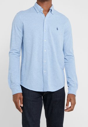 Homme portant une chemise bleu clair à manches longues avec un petit logo sur la poitrine et un jean foncé sur un fond uni.