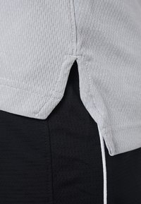 Jasnoszara sportowa koszulka z teksturowanym wzorem, z bocznymi rozcięciami i kontrastującą czarną tkaniną. Pokazuje gładki, oddychający materiał.