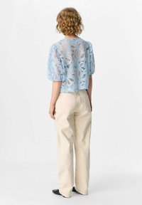 Woman with curly hair wearing sheer blue floral lace blouse, beige wide-leg pants, and black shoes, standing facing away on white background.