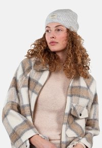 Gray knit beanie with a label, cream and brown plaid jacket with a zipper and chest pocket, and a beige sweater underneath.