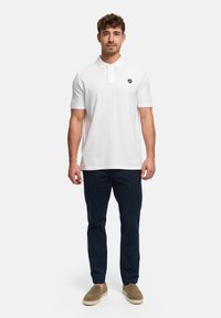Hvid poloshirt med krave og to knapper samt sort logo; kombineret med mørkeblå bukser og beige slip-on sko. Neutral tekstureret udseende.