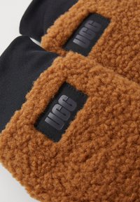 Guantes de forro polar texturizado marrón con un puño de tela negra y una etiqueta rectangular negra con "UGG" grabado en gris.