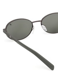Gafas de sol ovaladas con lentes verde oscuro, montura metálica fina y varillas verdes mate. Almohadillas nasales transparentes para mayor comodidad. Diseño minimalista.
