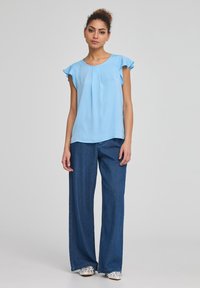Top azzurro chiaro a maniche corte con dettagli arricciati e accenti a volant, abbinato a pantaloni in denim blu a gamba larga e scarpe slip-on a fantasia.