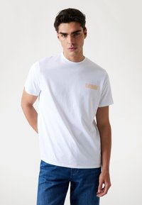 Witte katoen t-shirt met een ronde hals en een klein beige logo op de linkerkant van de borst, gecombineerd met blauwe denim jeans. Gladde textuur.