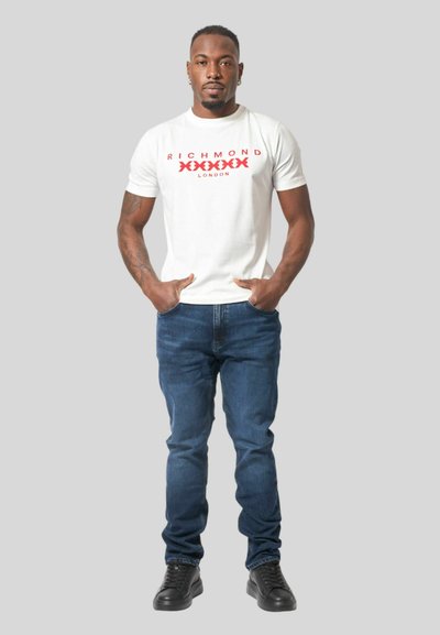 T-shirt bianca in cotone con testo rosso "RICHMOND LONDON", abbinata a jeans blu scuro e sneakers nere. La vestibilità è regolare con collo a giro.