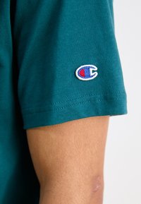 T-shirt de algodão em teal com mangas curtas e um logótipo bordado em branco, azul e vermelho na manga. Textura suave com um corte ajustado.