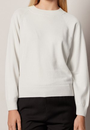 Femme portant un pull blanc uni à manches longues avec poignets et ourlet côtelés, assorti à un pantalon noir, vue du cou aux hanches.