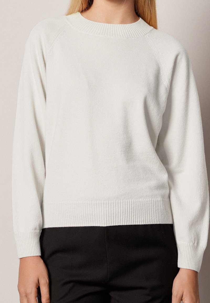 Femme portant un pull blanc uni à manches longues avec poignets et ourlet côtelés, assorti à un pantalon noir, vue du cou aux hanches.