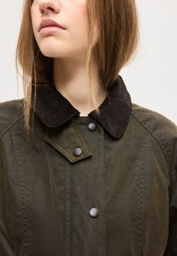 Zbliżenie na osobę ubraną w ciemnozieloną kurtkę z czarnym kołnierzem ze sztruksu i czarnymi zatrzaskami, patrzącą lekko w lewo.