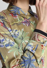 Blusa a maniche lunghe beige con un motivo floreale, caratterizzata da accenti verdi, blu e rossi. Colletto rigido e polsini con bottoni con bordi a contrasto.
