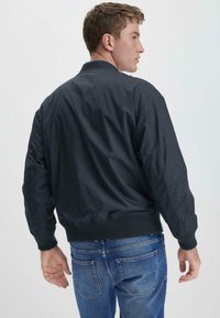 Dunkelmarineblaue Bomberjacke mit gerippten Bündchen und Saum, die eine glatte, leicht glänzende Textur aufweist. Normale Passform und keine sichtbaren Metallteile.