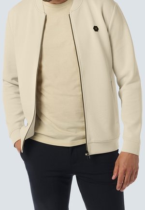 Mężczyzna w beżowej kurtce na zamek błyskawiczny, pod którą ma pasującą koszulkę, oraz w czarnych spodniach, z jedną ręką w kieszeni i widocznym częściowo tułowiem.