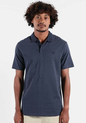 Homme aux cheveux bouclés portant un polo bleu marine à manches courtes et un pantalon de couleur claire, debout devant un fond blanc uni.