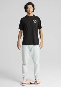 Sort T-shirt med korte ærmer og hvid "PUMA" logo, parret med lyseblå jeans og sneakers med orange accenter mod en neutral baggrund.