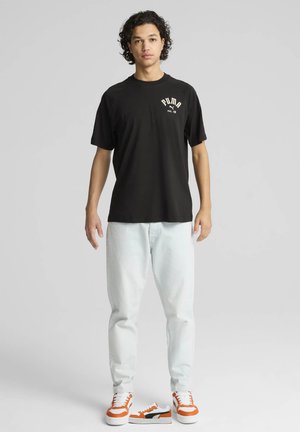 Sort T-shirt med korte ærmer og hvid "PUMA" logo, parret med lyseblå jeans og sneakers med orange accenter mod en neutral baggrund.