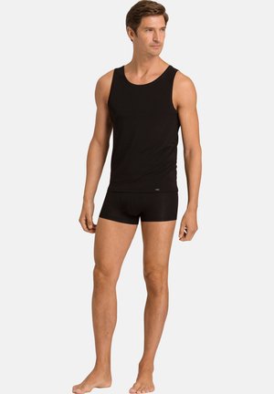 Mand iført sort ærmeløs undertrøje og sorte boxer shorts, stående barfodet mod en ensartet lys baggrund, kiggende mod højre.