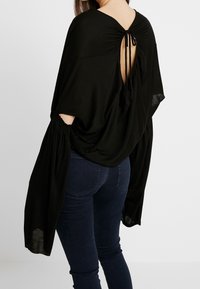 Haut noir ample avec manches chauve-souris, doté d'une fermeture nouée à l'arrière et d'un tissu texturé. Associé à un jean bleu foncé.