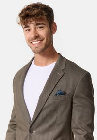 Blazer marrone a scacchi con rever a punta, dotato di un fazzoletto da taschino blu a fantasia. T-shirt bianca sottostante, con texture di tessuto morbido.