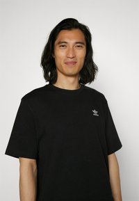 Čierne bavlnené tričko s krátkym rukávom a okrúhlym výstrihom. Na ľavej strane hrudníka má malé biele logo Adidas.