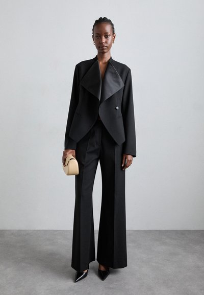 Sort skræddersyet blazer med bred krave, parret med matchende flagrende bukser. Modellen holder en lille beige clutch og har spidse sorte hæle på.