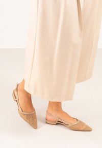 Zapatos slingback beige con punta afilada, que presentan un patrón similar a una malla en la parte frontal y un tacón plano, mostrados con pantalones holgados color crema.