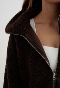 Bruine fleece jas met een rits aan de voorkant, grote kraag en een gestructureerd oppervlak. Draagt over een witte top, met de nadruk op de halslijn en het ritsdetail.