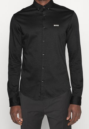 Uomo che indossa una camicia nera aderente con bottoni e piccolo logo "BOSS" sul petto sinistro, abbinata a pantaloni scuri.