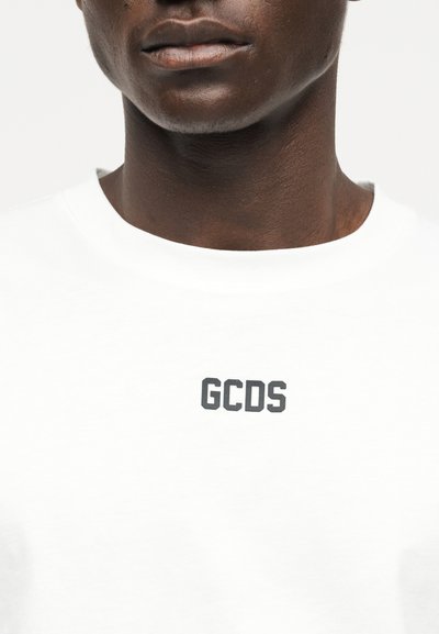 Κοντινό πλάνο ατόμου που φοράει λευκό T-shirt με το "GCDS" τυπωμένο μαύρο στο στήθος.