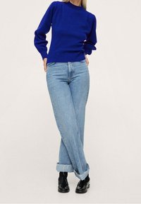 Pull bleu tricoté à manches longues associé à un jean bleu clair ample, revers remontés, et chaussures noires. Design et texture simples.