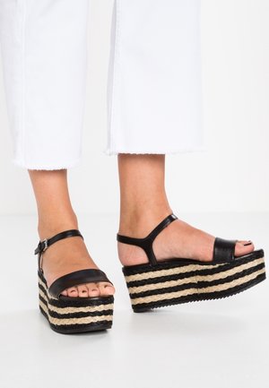 Füße in schwarzen Plateausandalen mit gestreiften, gewebten Sohlen und Knöchelriemen, kombiniert mit weißen, gekürzten Hosen mit ausgefransten Säumen.
