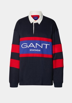 Tumesinise pikkade varrukatega ragbipait, millel on valge krae, punased ja sinised horisontaalsed triibud ning suur valge "GANT SPORTSWEAR" logo.