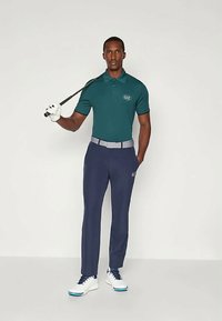 Τιρκουάζ πόλο μπλούζα με λογότυπο, γκρίζι ζώνη, ναυτικές παντελόνες και λευκά παπούτσια γκολφ. Η φιγούρα κρατά ένα μπαστούνι γκολφ. Απλός σχεδιασμός και μοντέρνα εφαρμογή.