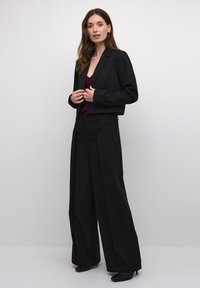 Crni skraćeni sako od strukturirane tkanine, uparen s hlačama širokog kroja visokog struka. Outfit sadrži tamnocrveni top ispod.