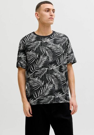 Ung mand iført en sort og hvid T-shirt med palmebladsmønster og korte ærmer samt sorte bukser, stående foran en ensfarvet lys baggrund.