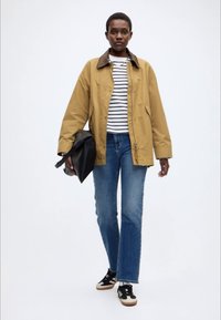 Veste légère kaki avec col marron, chemise rayée, jean bleu et baskets noires. Modèle tenant un sac noir, debout devant un fond uni.