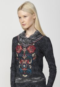 Schwarz gemusterte Langarmbluse mit buntem Blumenstich in Rot, Blau und Gold; Schalkragen mit strukturiertem grauem Besatz.
