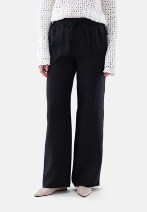 Frau trägt schwarze, weit geschnittene Hosen mit Kordelzug, weißen, offen gestrickten Pullover und beige spitze Ballerinas, steht vor weißem Hintergrund.
