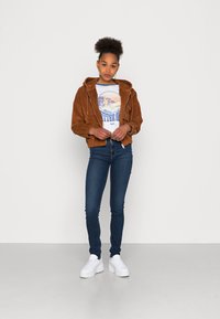 Casaco de cotim castanho com capuz, ajustado na cintura, usado sobre uma t-shirt gráfica clara, combinado com jeans skinny escuros e ténis brancos.